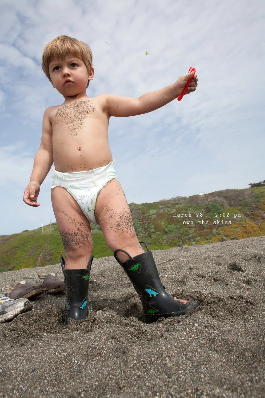 A toddler in a diaper and rain boots stands on a sandy hill, captioned own the skies.
