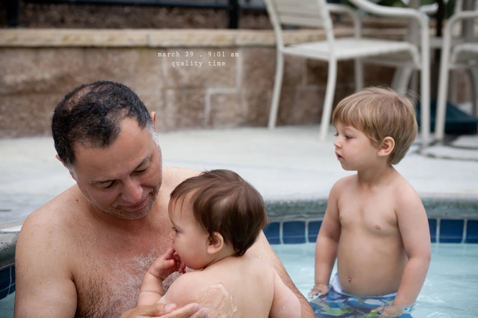 A man holds a baby in a hot tub beside a toddler, captioned quality time.