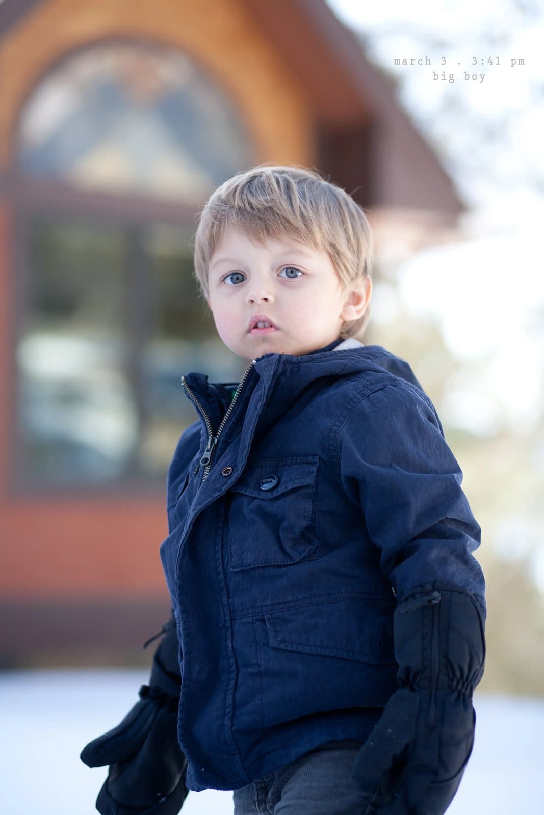 A toddler in a navy jacket and mittens stands outside a house, captioned big boy.