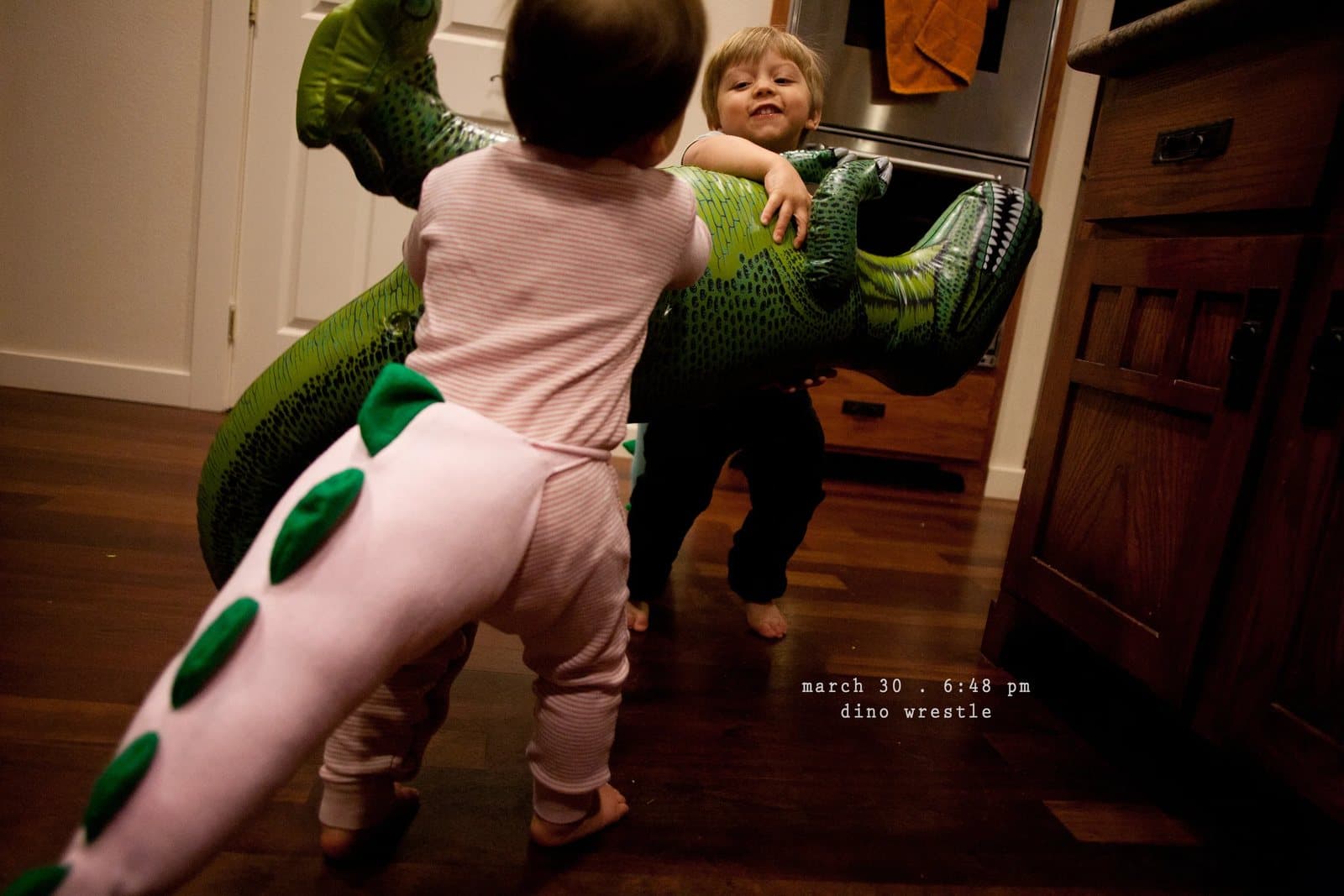 A baby in a dinosaur tail costume wrestles an inflatable dinosaur, captioned dino wrestle.
