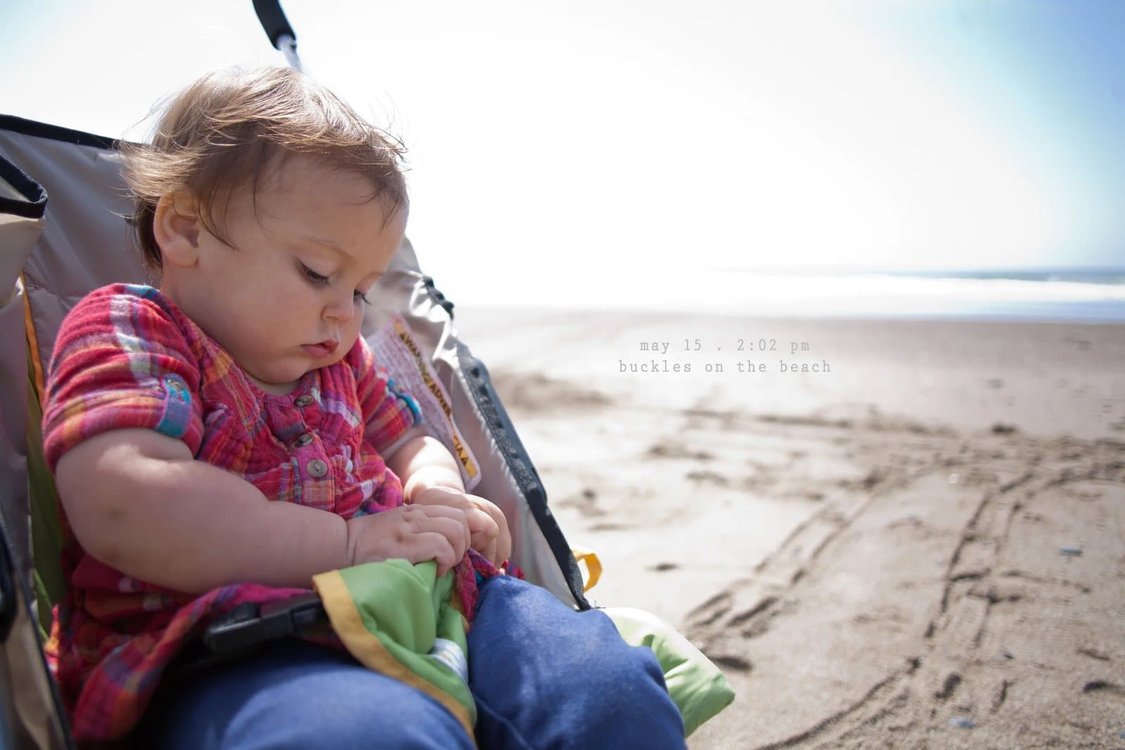A baby in a plaid shirt sits in a stroller on a beach, captioned buckles on the beach.