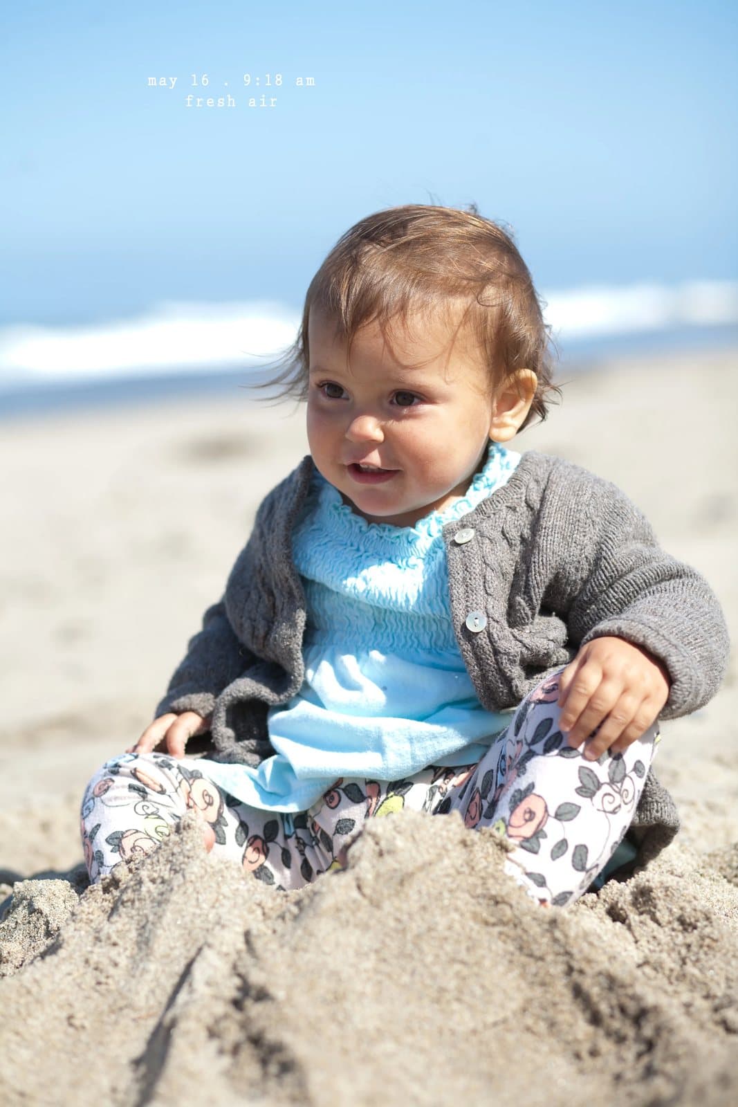 A smiling baby in a gray cardigan sits in the sand at the beach, captioned fresh air.