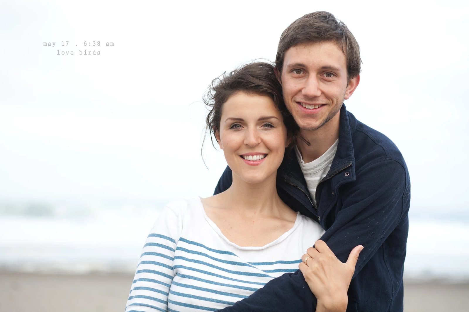 A smiling couple embraces on a beach, captioned love birds.