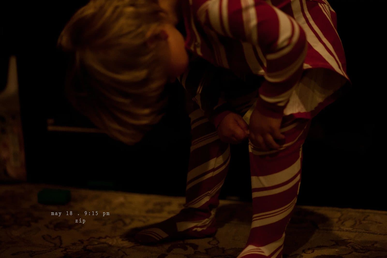 A child in striped pajamas bends to zip up in a dark room, captioned zip.