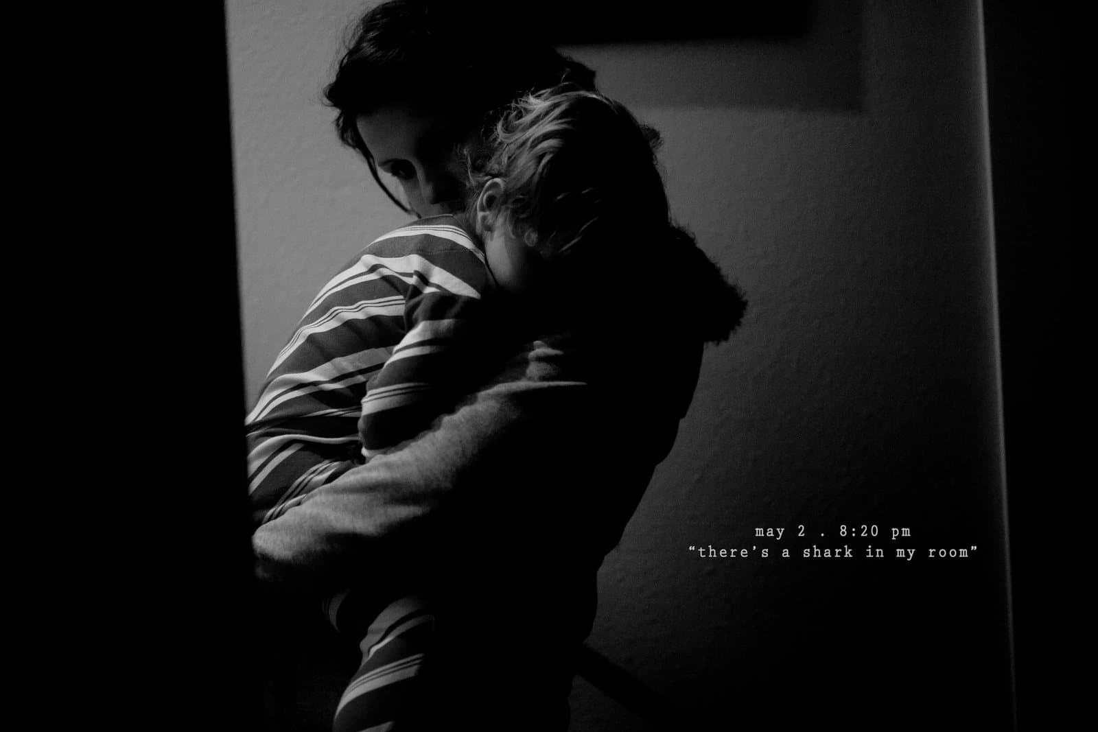 A woman holds a child in the dark by a doorway, captioned there's a shark in my room.