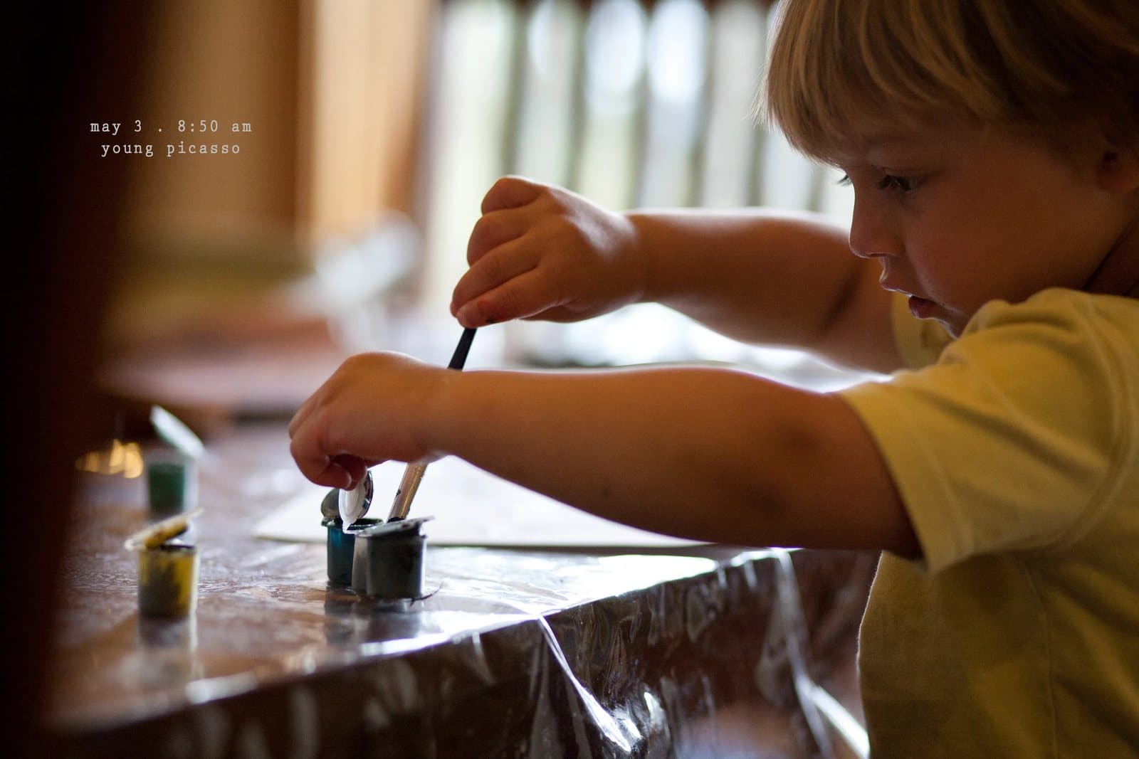 A boy dips a paintbrush into small paint pots at a table, captioned young picasso.
