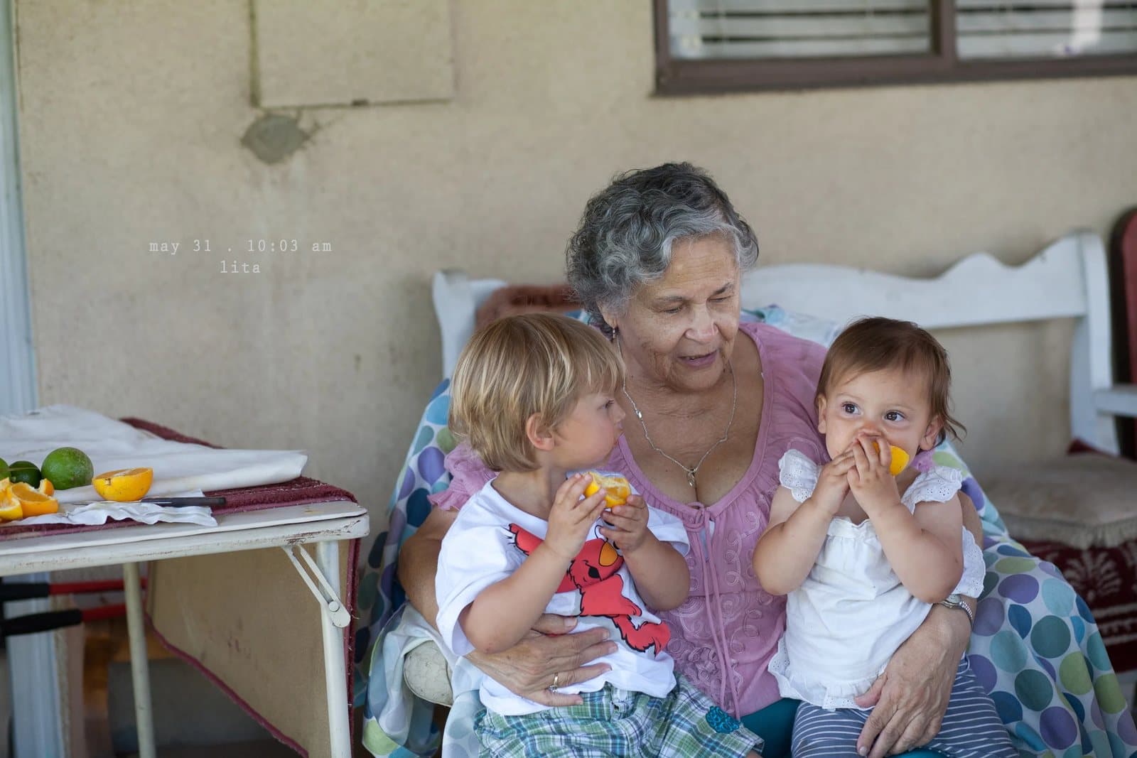 A gray-haired woman sits on a porch with a toddler and a baby eating fruit.