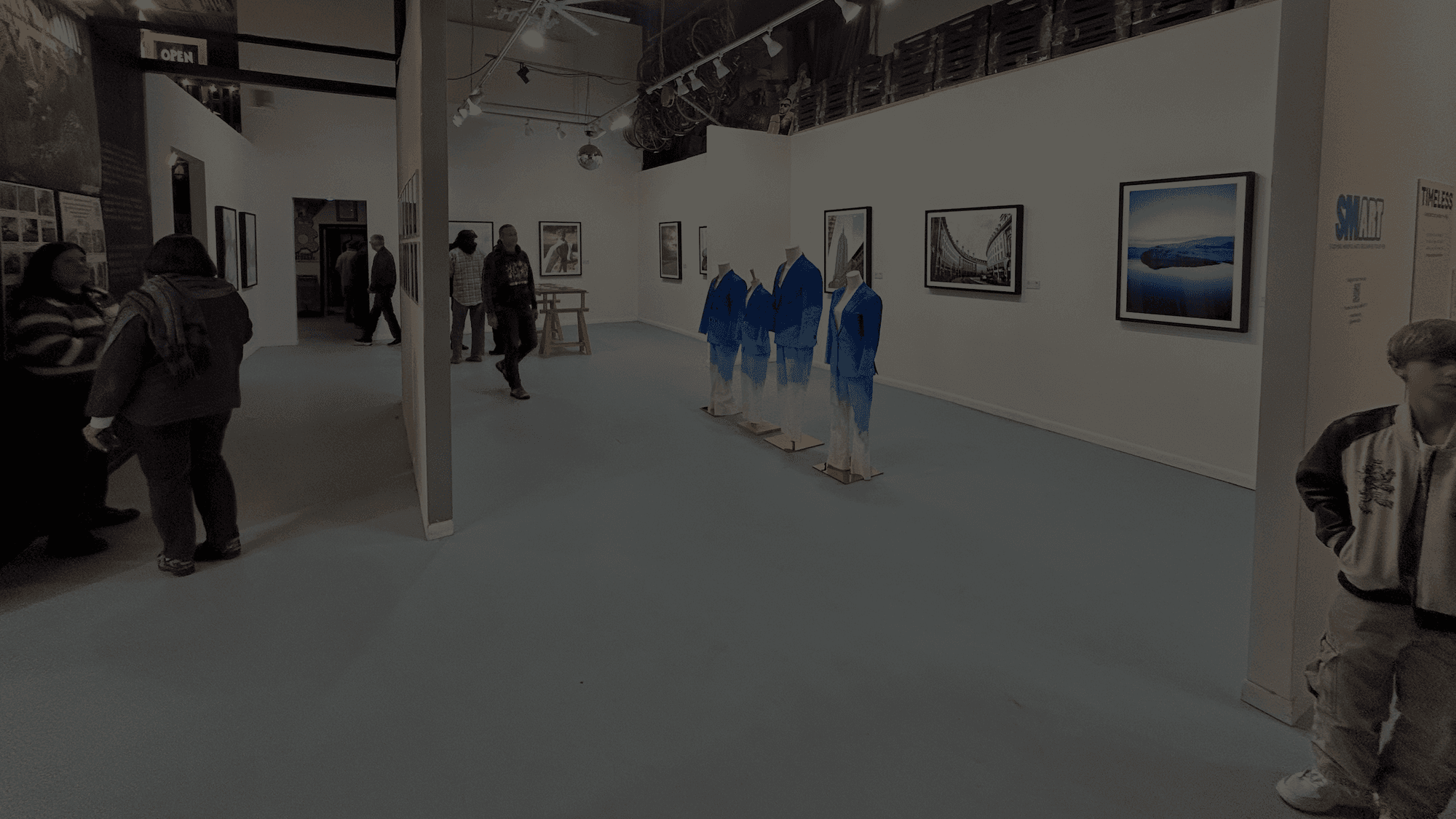 Visitors walk through the TIMELESS exhibit at 1906 Gallery, with painted blue suits on mannequins among framed photographs.