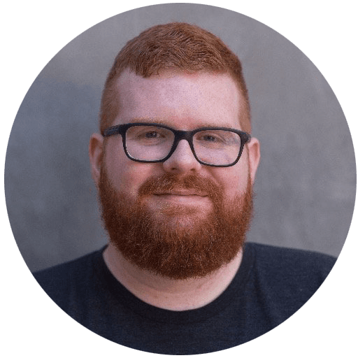 Portrait of Kyle Adams, bearded and in glasses, smiling against a gray concrete wall.