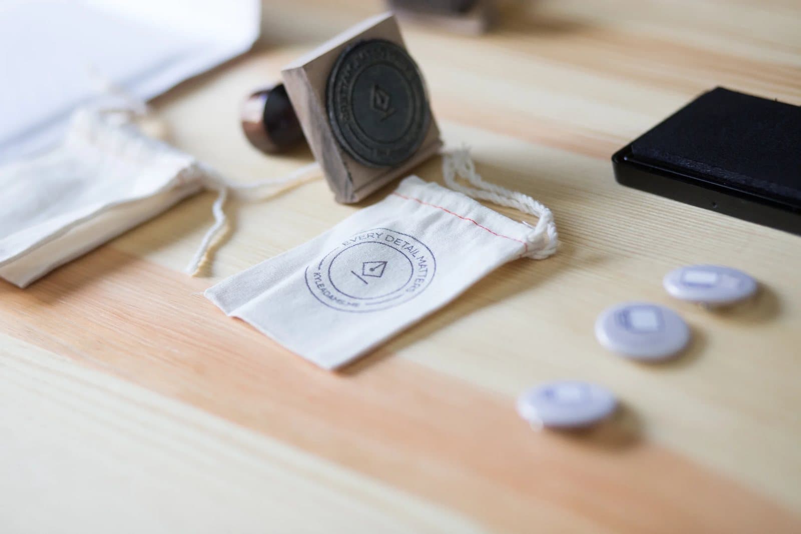 Branded cotton pouches, a rubber stamp, and small buttons from Kyle's brand on a pine desk.