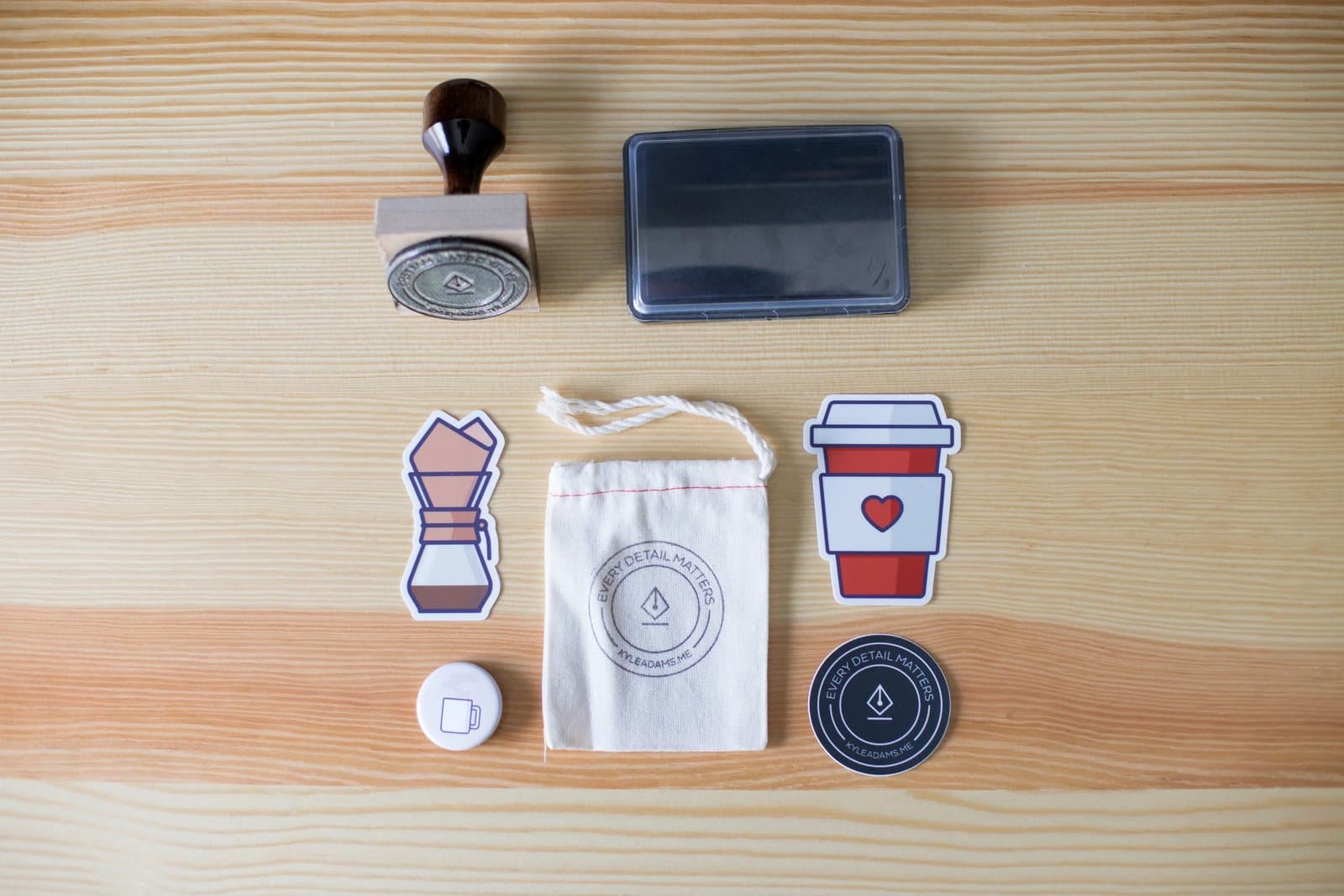 Flat lay of Kyle's coffee icon stickers, a branded pouch, button, stamp, and ink pad on pine.
