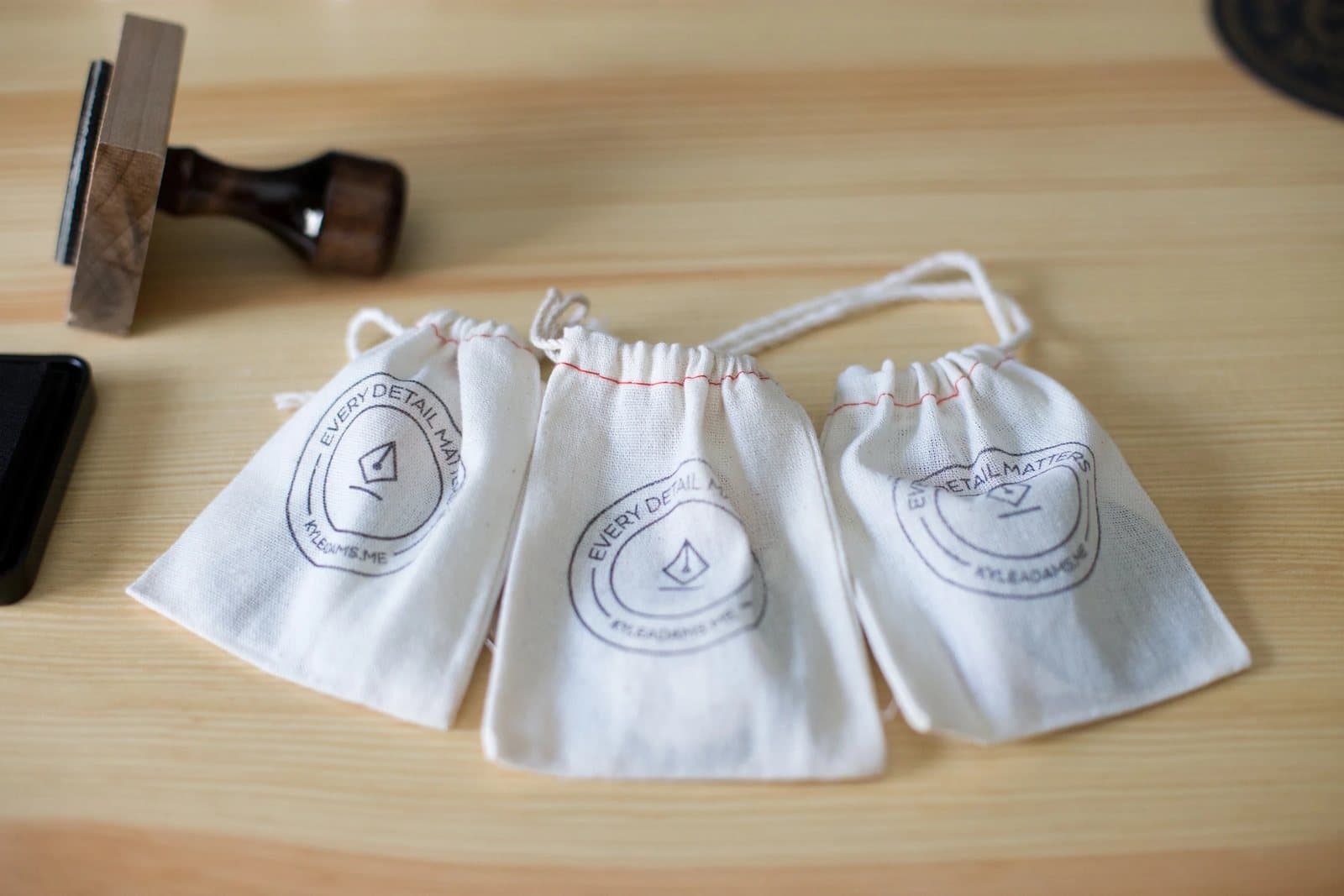 Three drawstring pouches stamped with Kyle's Every Detail Matters seal lie on a light wood desk.
