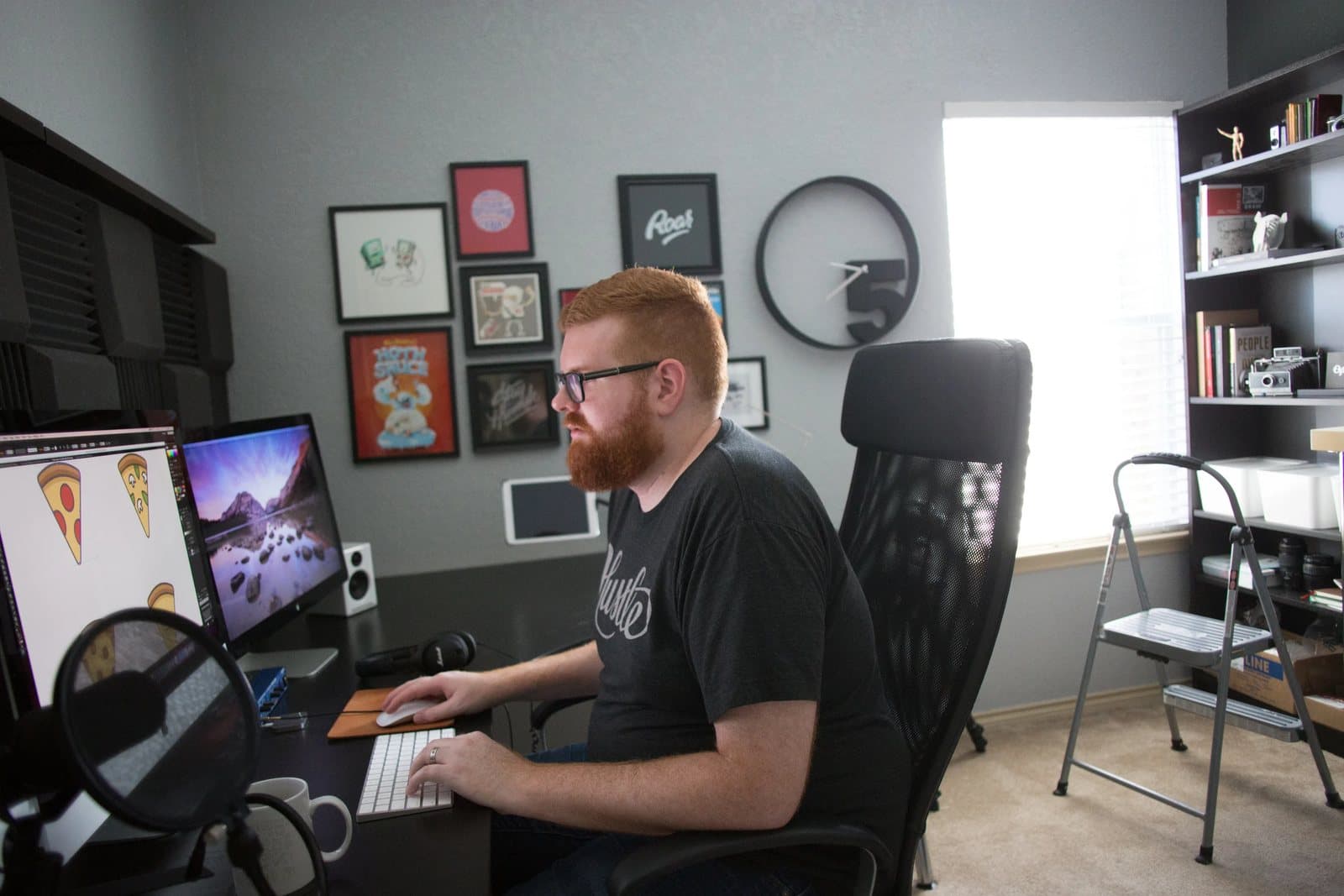 Kyle works at his desktop computer in a studio lined with framed prints and shelves.