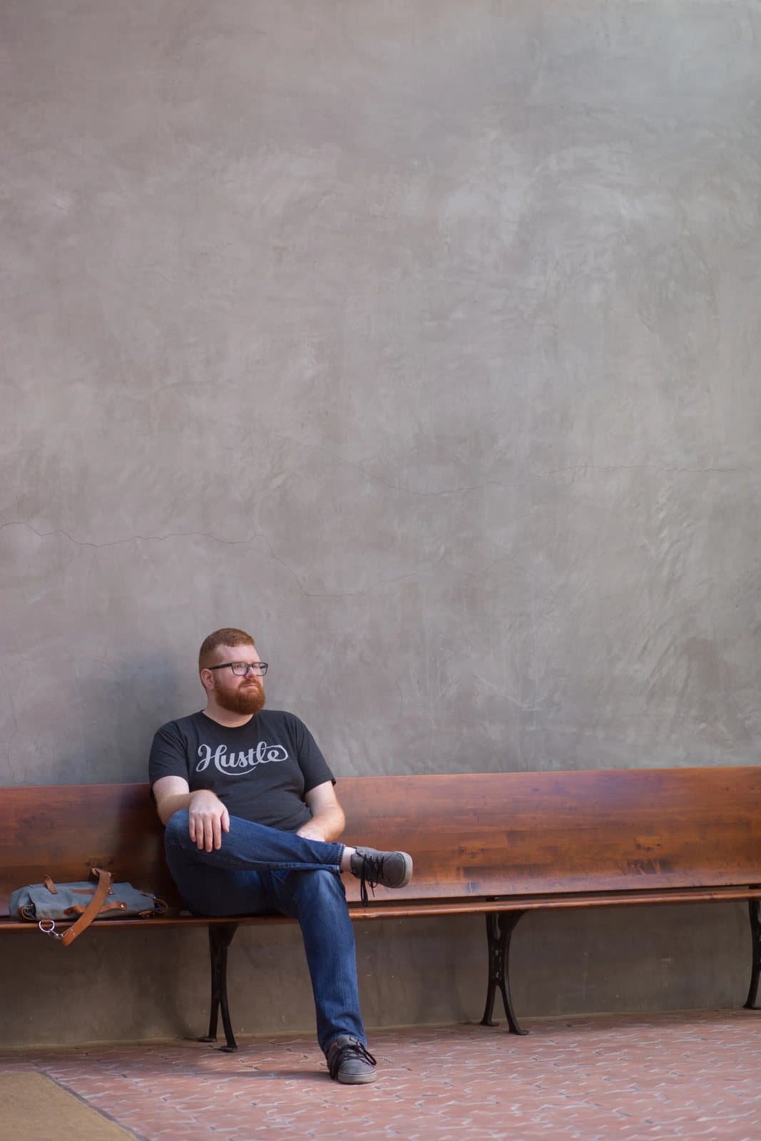 Kyle sits on a wooden bench against a tall plaster wall, looking off to the side.
