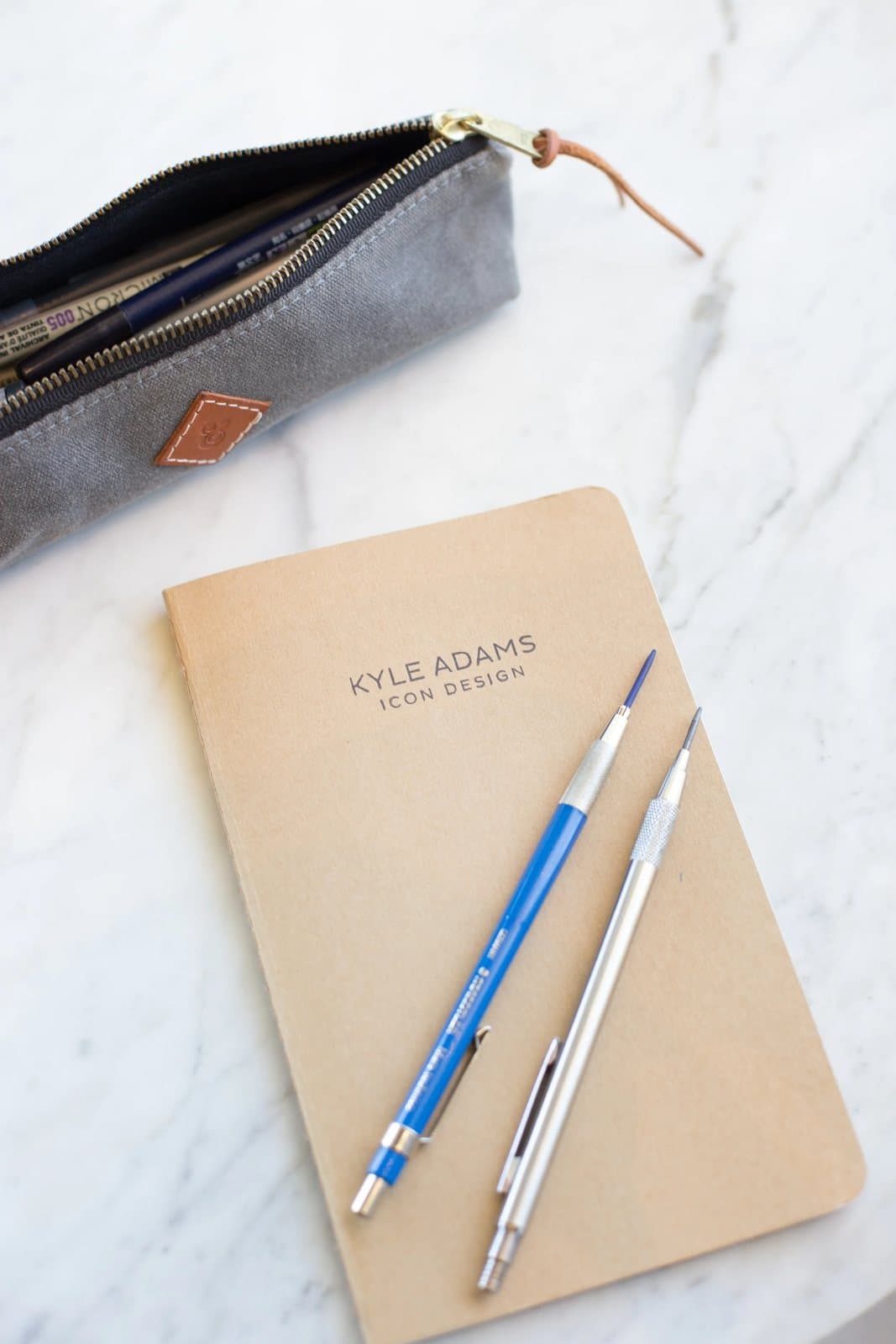 A notebook reading Kyle Adams Icon Design with pencils and a pencil case on white marble.