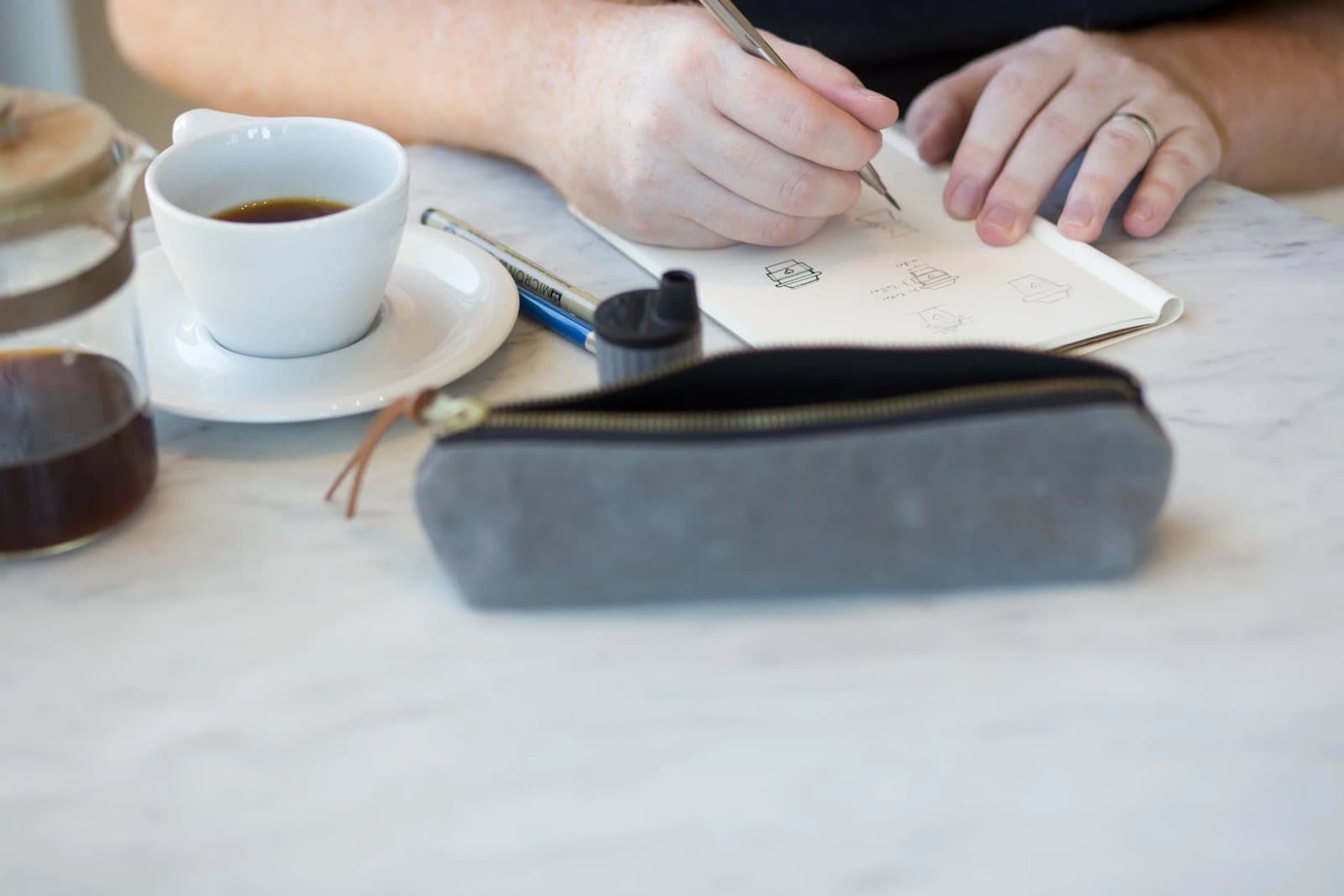 Kyle sketches icons in a notebook beside his coffee and gray pencil case at the cafe.