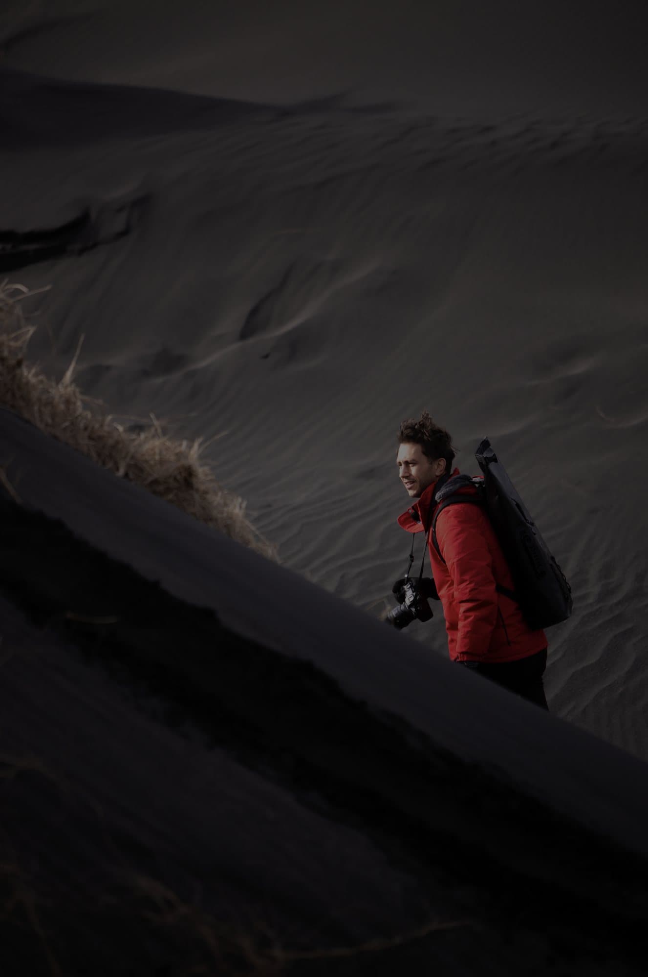 Mark with a camera and red jacket climbing a black sand dune in dim light.