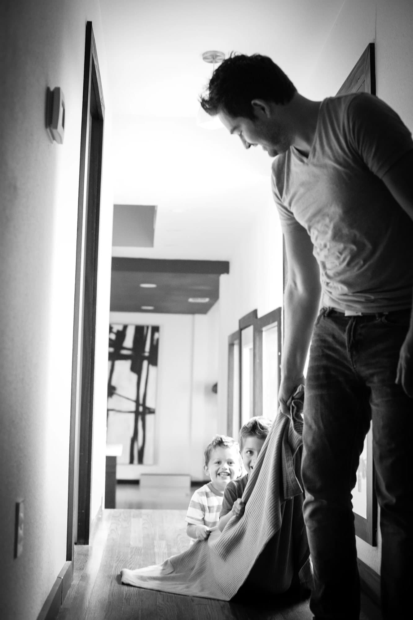 Black and white shot of dad pulling the grinning boys down the hall on a blanket.