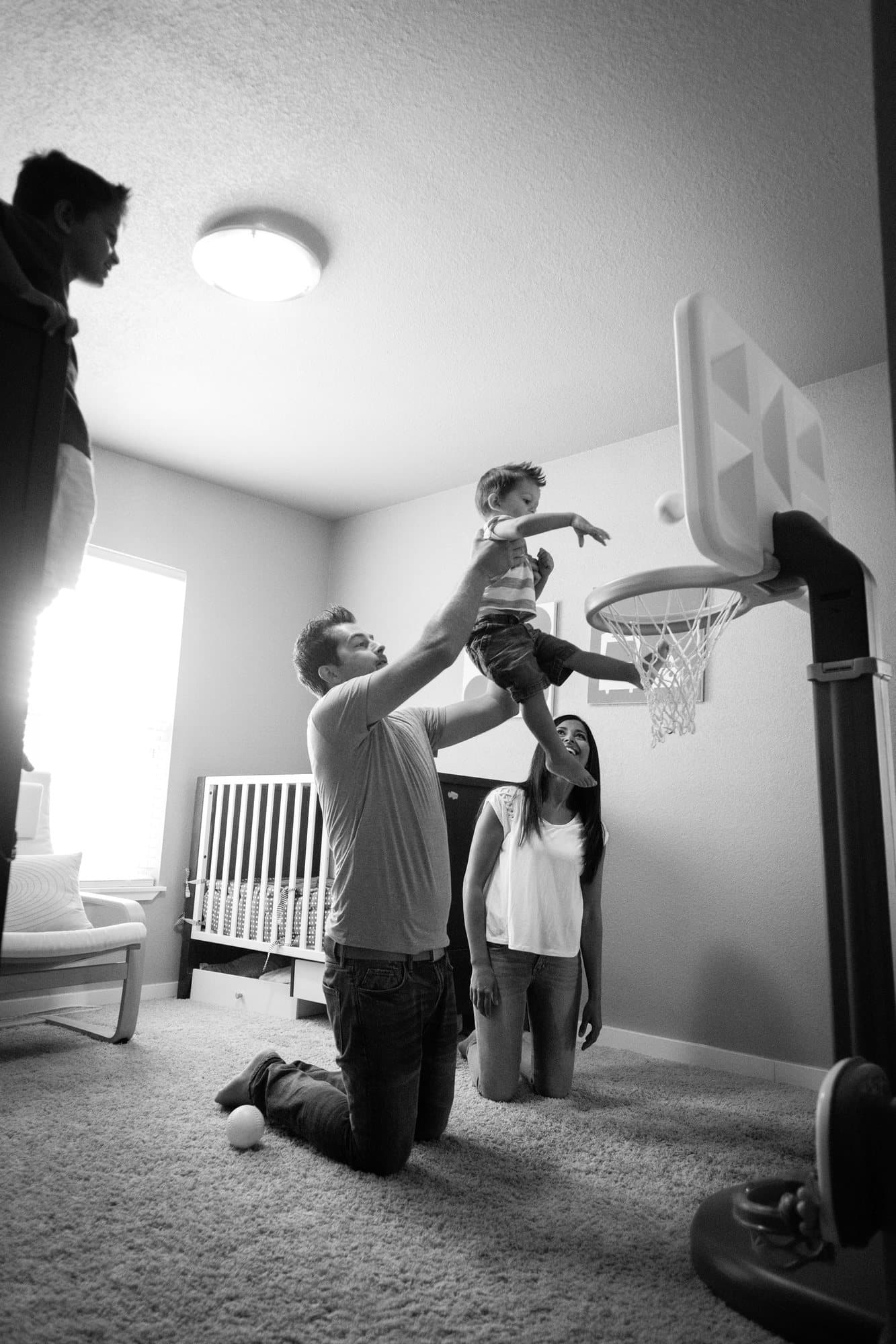Black and white shot of dad lifting a toddler to dunk on a toy hoop as mom watches.