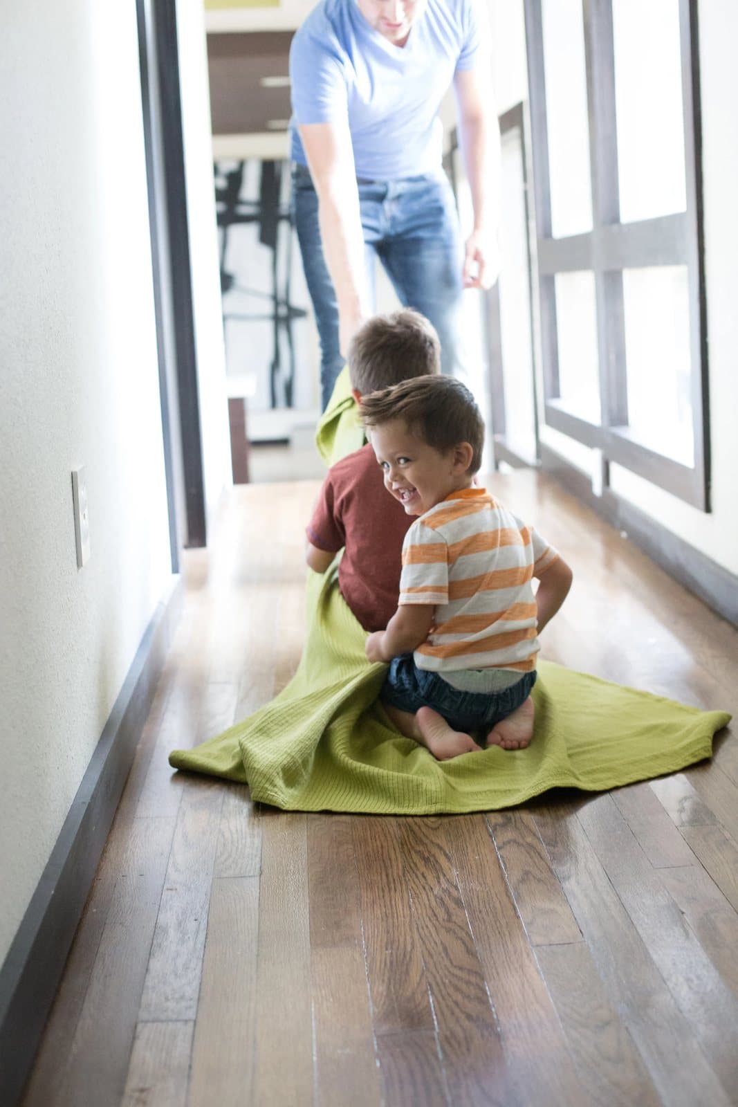 The boys sit on a green blanket in the hallway, one grinning back, as dad walks behind.