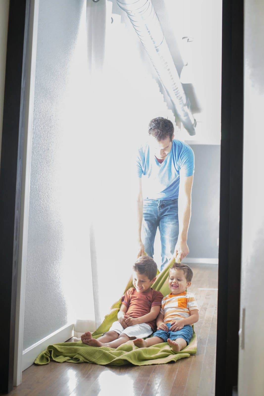 Dad pulls both boys along the glowing hallway floor on a green blanket, a family ritual.