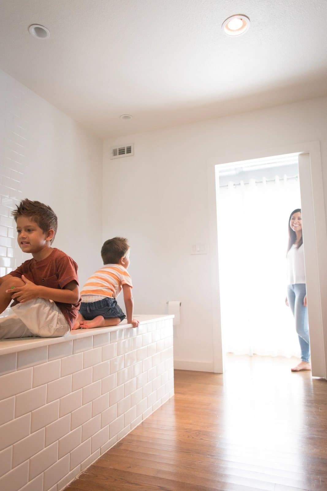 The boys perch on a white tiled tub ledge while mom smiles from the bathroom doorway.