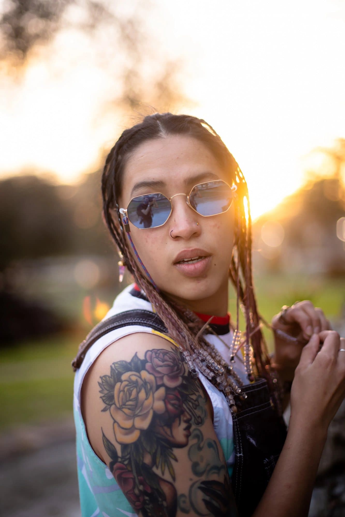 Elena Carrasco, featured in Volume 35, in round sunglasses with braids and a floral arm tattoo at sunset.