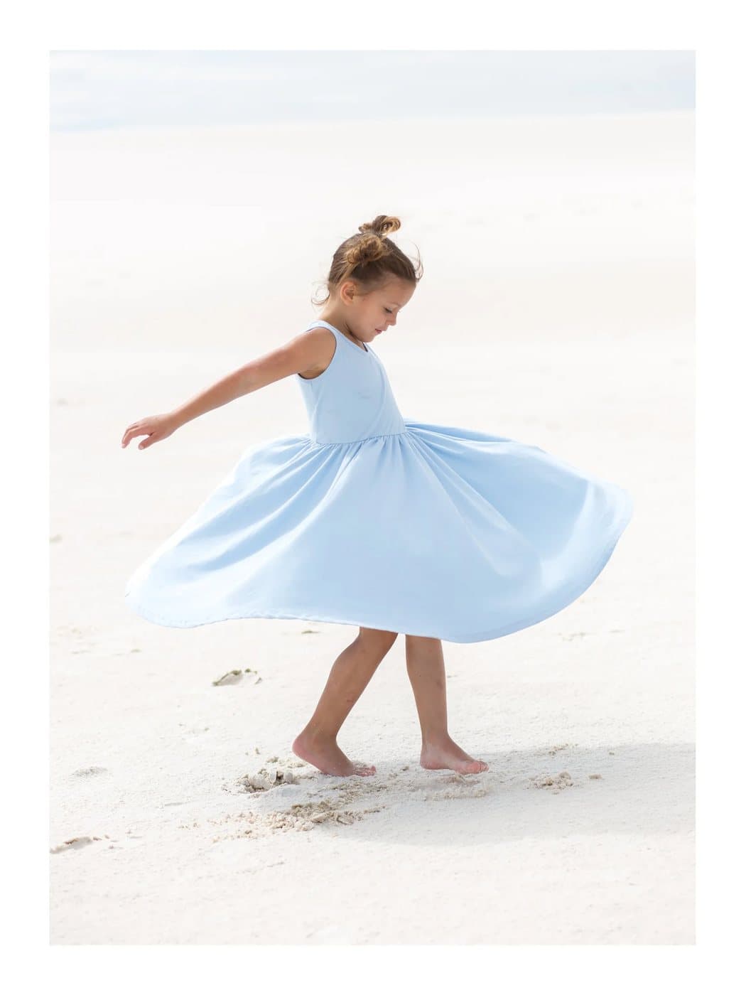 Aila, a TIMELESS archival print of a young girl twirling in a pale blue dress on a white beach.