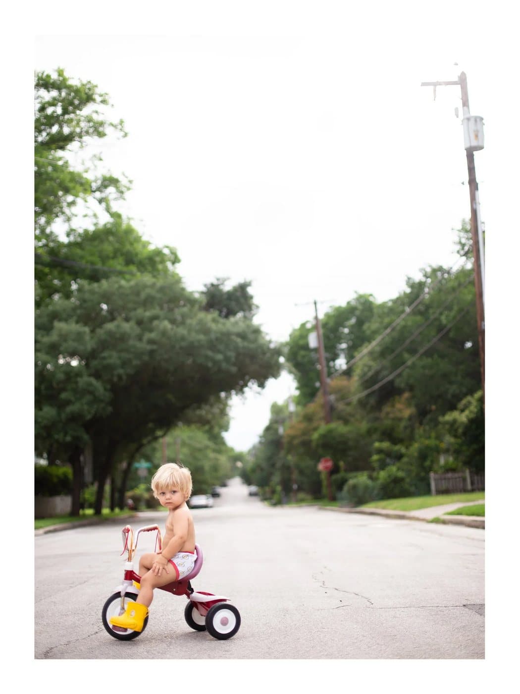 Luca, a TIMELESS archival print of a toddler in yellow boots on a red tricycle in a leafy street.