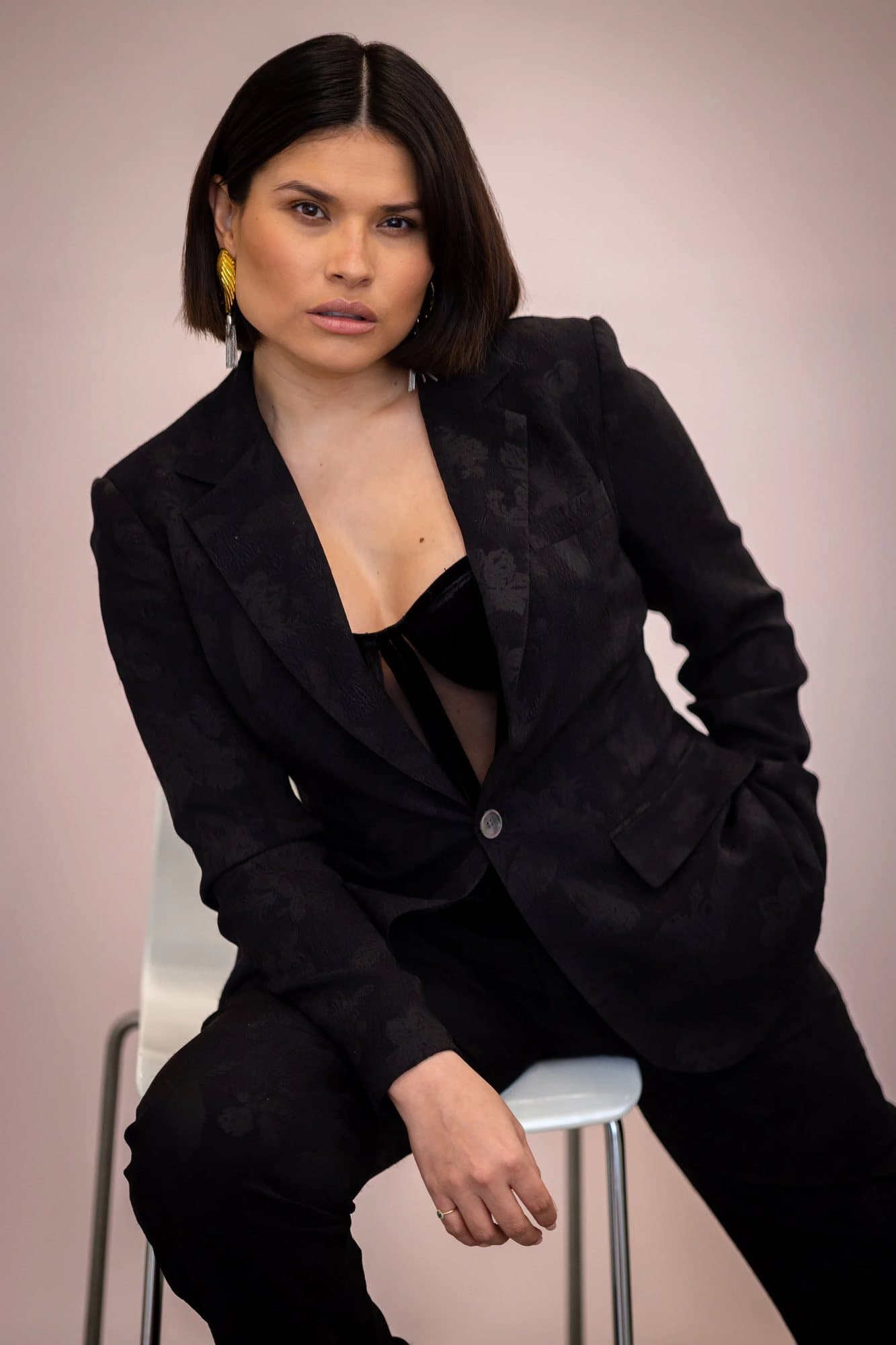 Grecia Valencia in a black jacquard suit and gold earrings, perched on a white stool.