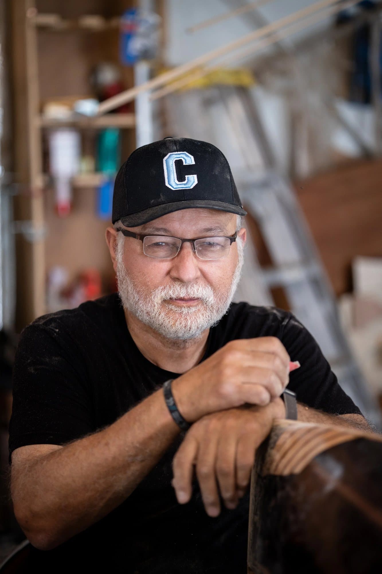 Carlos Becerra in glasses and a black C cap, leaning on a chair in his workshop.