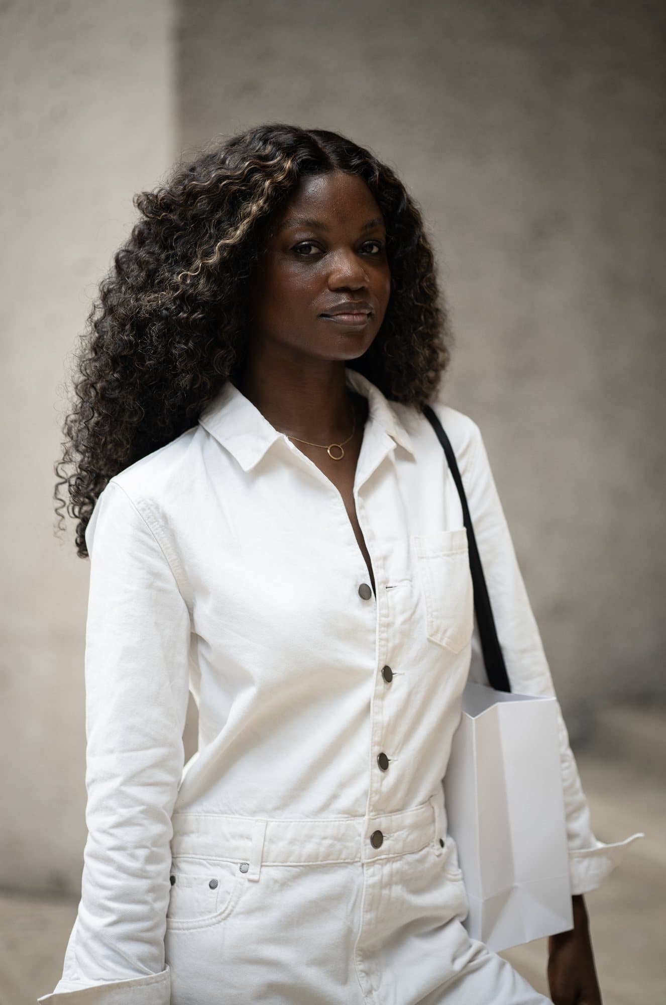 Zola-Ludivine Luvualo in a white jumpsuit, carrying a white shopping bag against a concrete wall.
